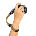 A hand holding a camera attached to a brown camera strap with black adjustment elements.