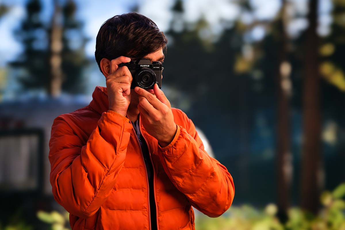 Person in an orange jacket holding a camera outdoors with trees in the background