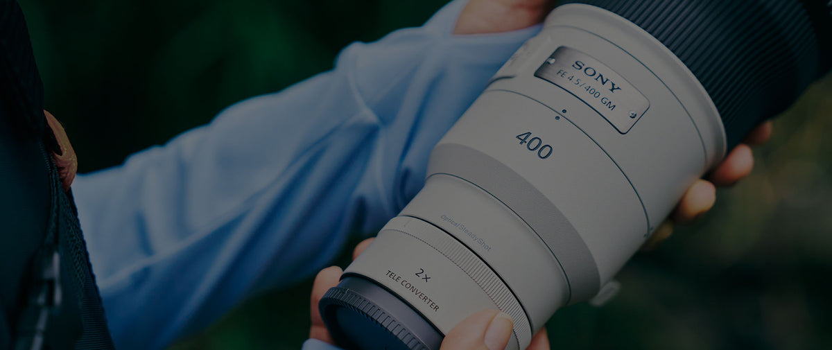 A close-up of a Sony 400mm f/2.8 lens held by a person wearing a light blue shirt, with a dark, blurred outdoor background.