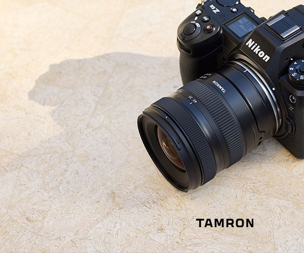 A Nikon full-frame camera with a Tamron lens resting on a textured beige stone surface, with the brand name 'TAMRON' printed in black text at the bottom right.