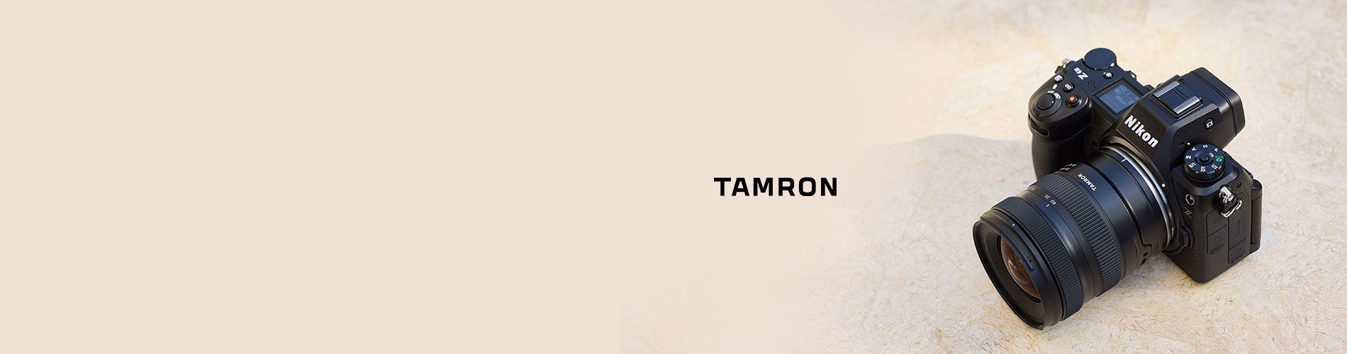A Tamron camera lens and camera body on a light beige textured background with the brand name 'TAMRON' printed in the center.