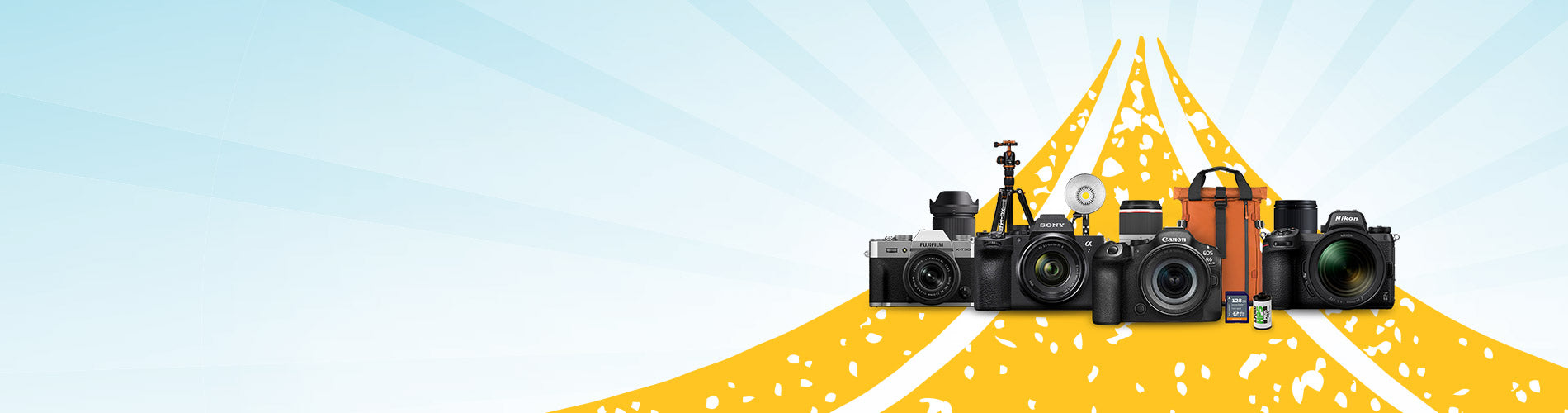 A group of cameras, lenses, and camera accessories arranged on a stylized yellow road with white dashed lines against a light blue background with sunburst rays.