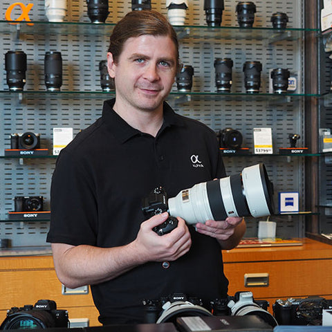 Man holding a camera lens in a store with camera equipment on shelves.