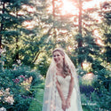 Bride in a white dress with a veil standing in a forest with sunlight filtering through the trees.