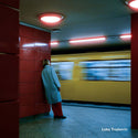 Person in a blue coat leaning against a wall in a subway station with a blurred train in the background.