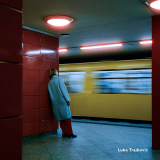 Person in a blue coat leaning against a wall in a subway station with a blurred train in the background.