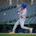 A baseball player in a gray uniform and blue helmet swings a bat at a baseball on a field, with stadium seats blurred in the background.