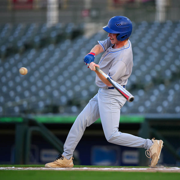 A baseball player in a gray uniform and blue helmet swings a bat at a baseball on a field, with stadium seats blurred in the background.
