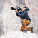 A man in a dark puffer jacket, tan pants, and a green cap kneels on a rock, holding a large telephoto camera with a long lens against a blurred rocky background.