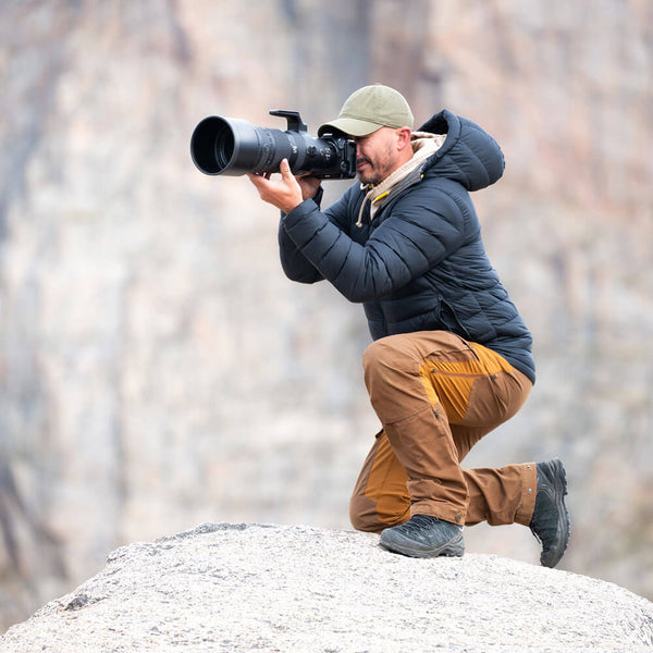 A man in a dark puffer jacket, tan pants, and a green cap kneels on a rock, holding a large telephoto camera with a long lens against a blurred rocky background.