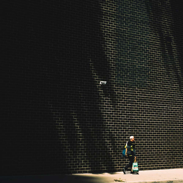 A person walking along a sidewalk beside a dark brick wall, with dramatic shadows cast across the wall and pavement.